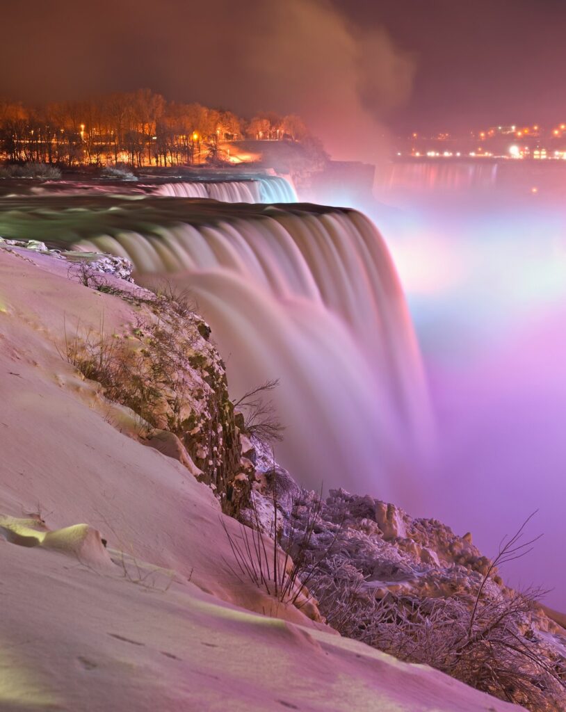 The American Falls lit in pink and blue at night, seen from Prospect Point with snow on the bank