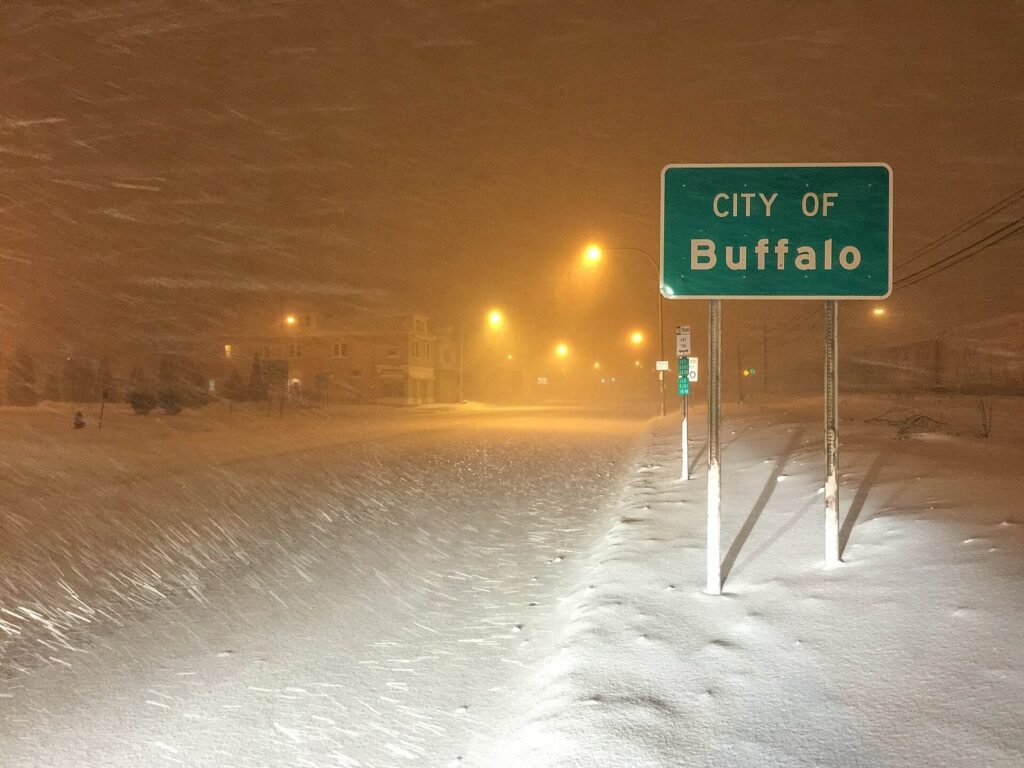 A green City of Buffalo sign half buried in blowing snow under orange streetlights during a lake-effect storm at night