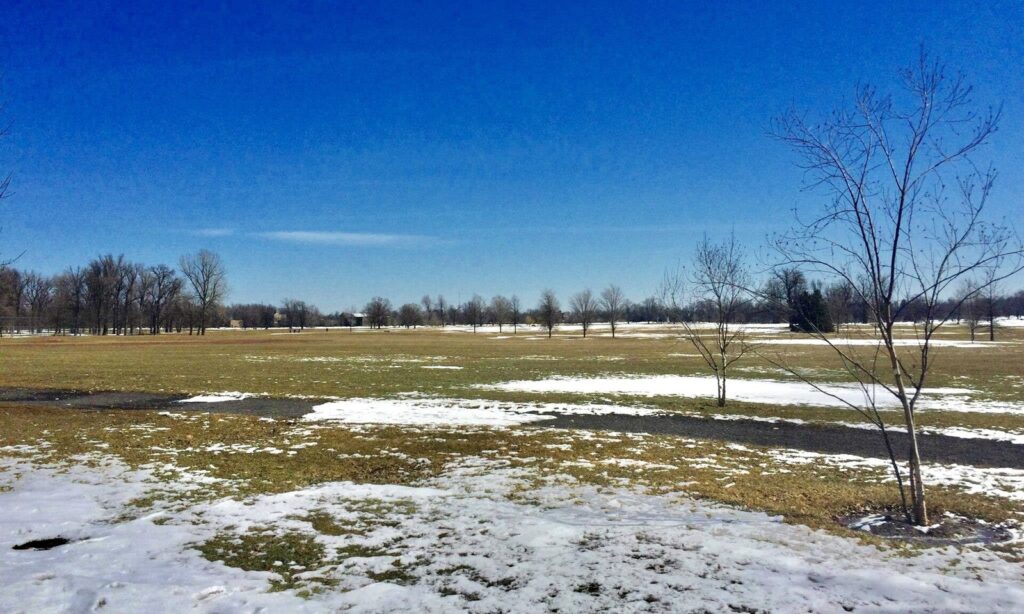 Patches of late snow on the brown grass of the Meadow in Delaware Park under a bright blue spring sky