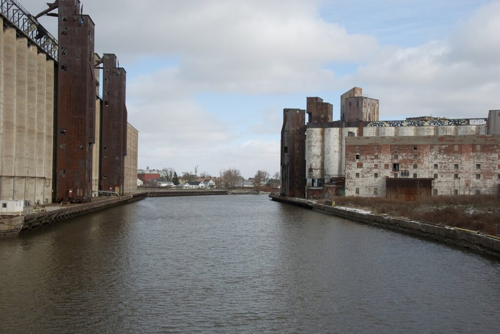 Tall concrete grain elevators lining both banks of the Buffalo River under a cloudy sky