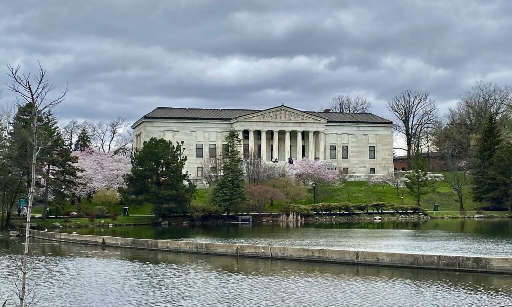 The Buffalo History Museum's marble portico above Mirror Lake, with pink cherry trees blooming in the Japanese Garden, a highlight of Buffalo NY spring things to do