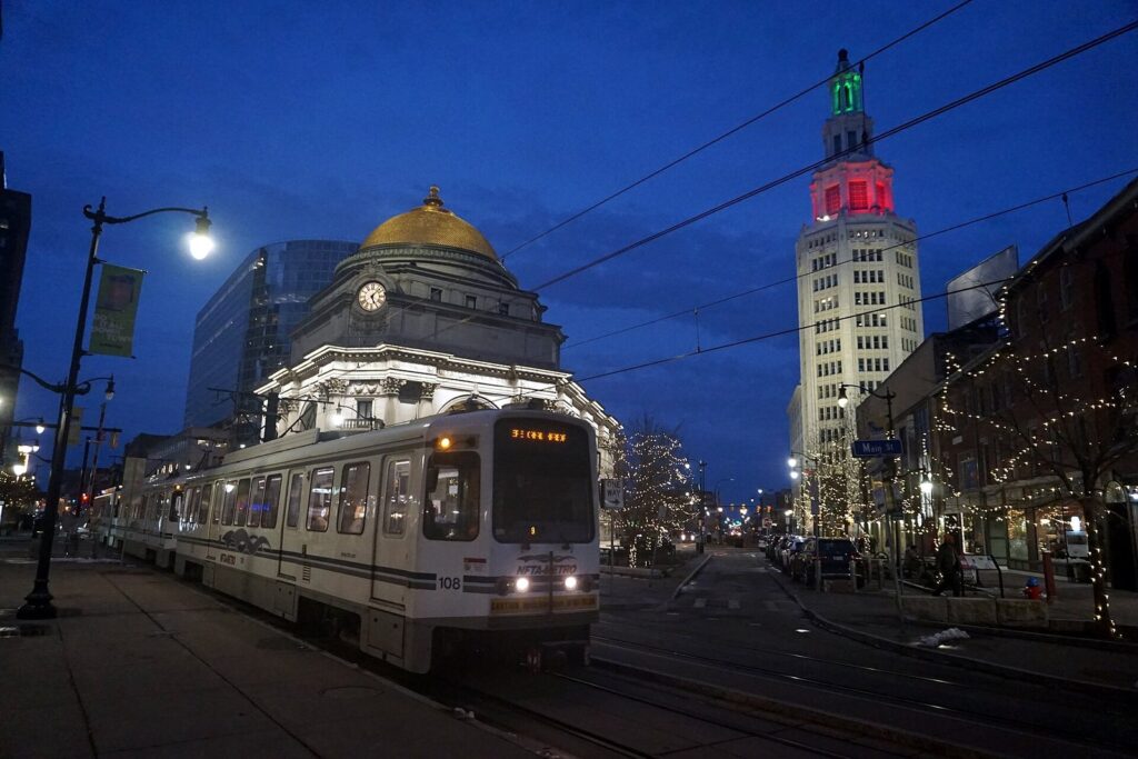 An NFTA Metro Rail train on Main Street in downtown Buffalo at dusk, with the gold dome of the old Buffalo Savings Bank and the lit Electric Tower behind