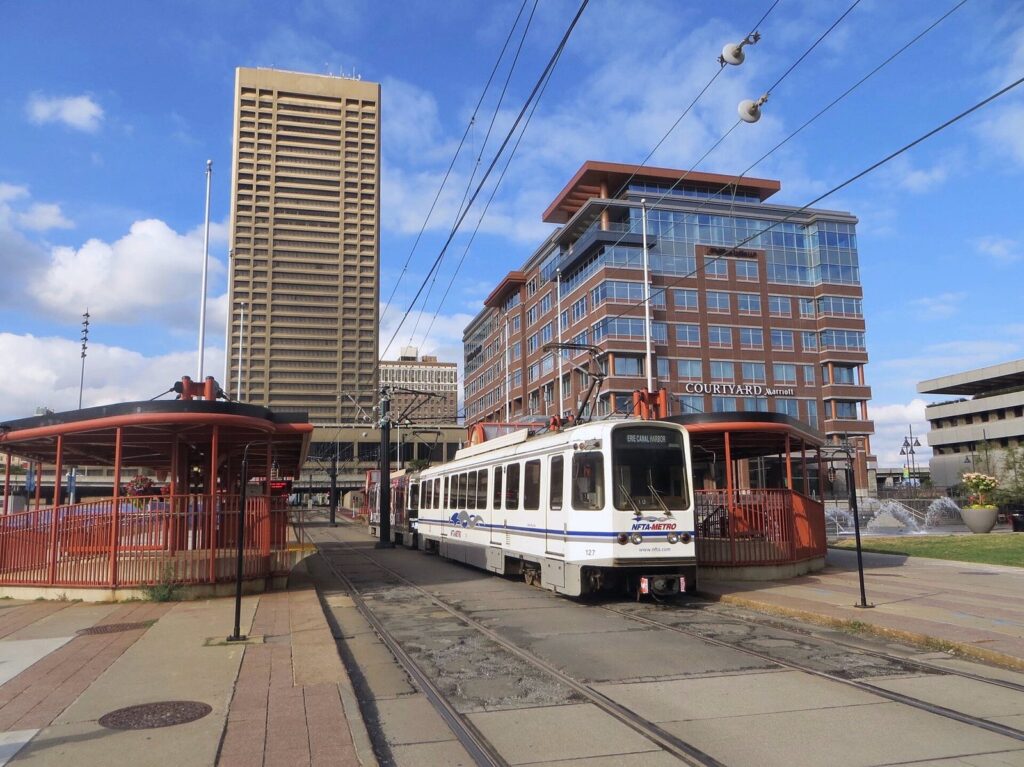 A white NFTA Metro Rail train at the Erie Canal Harbor station at Canalside, with downtown office towers behind