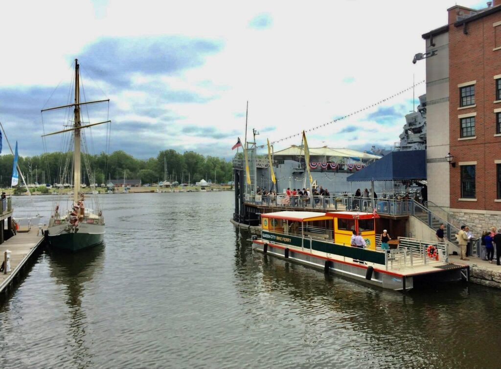 The Queen City Bike Ferry docked at Canalside's Commercial Slip beside a tall ship on a summer day, one of the easiest Buffalo NY summer things to do
