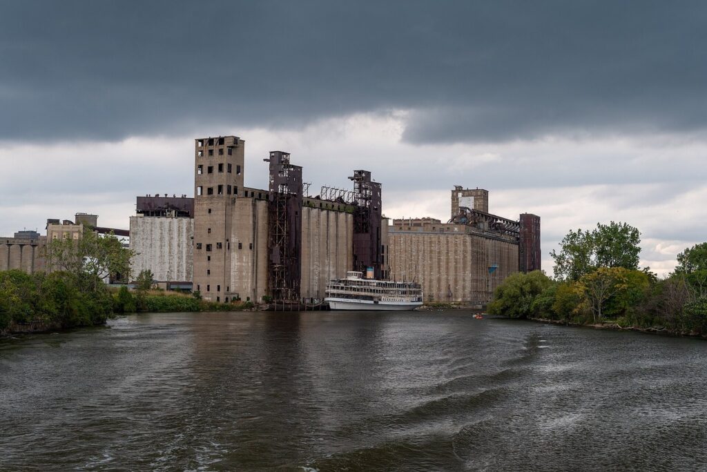 Massive concrete grain elevators of Silo City rising above the Buffalo River under a stormy sky, with a white steamship docked in front
