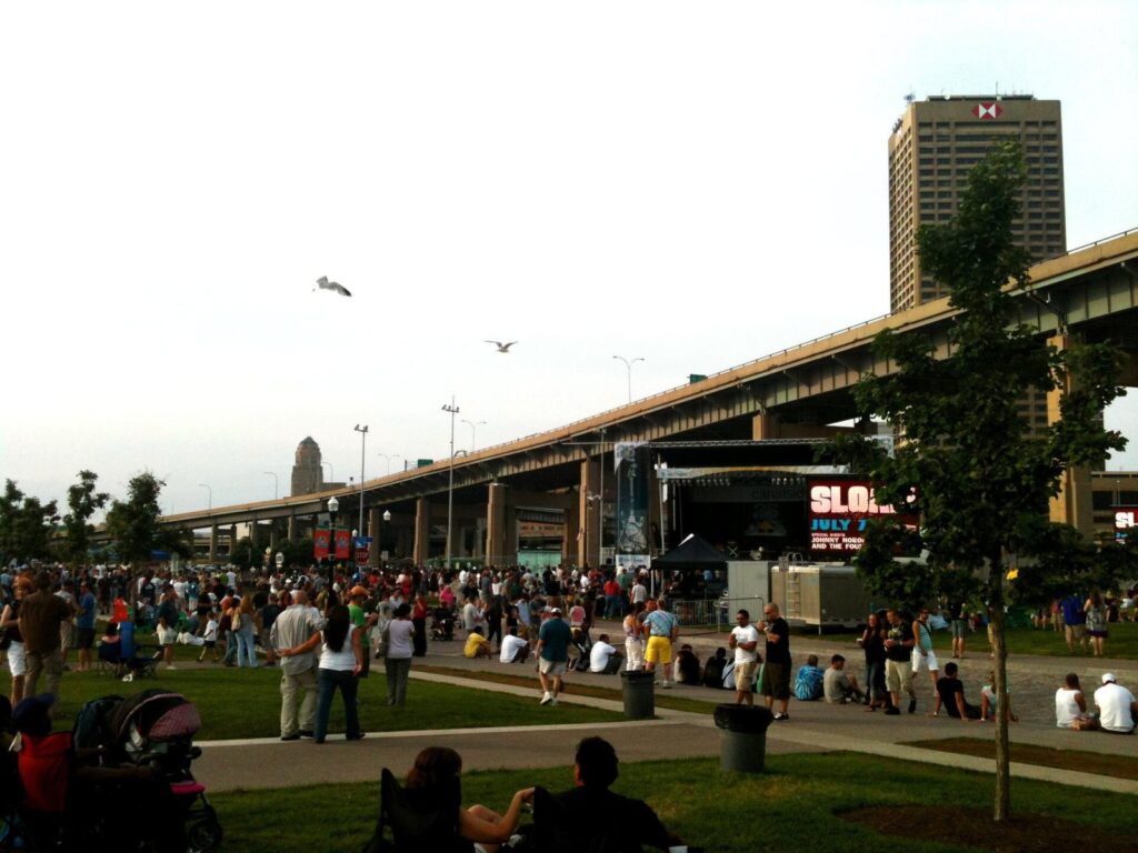 A crowd on the lawn at a summer concert on Buffalo's waterfront, with the Skyway and downtown towers behind
